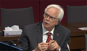 A man in a suit holds his hands in front of him as he speaks in a meeting.