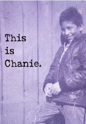 A black and white photo of a little boy shows him leaning against a wood structure wearing a jacket. He is smiling shyly toward the camera.