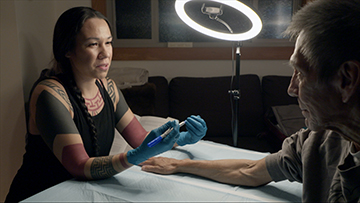 A bright halo light shines down on the outstretched arm of a man. He reached across a table to a woman who is tatooing him.