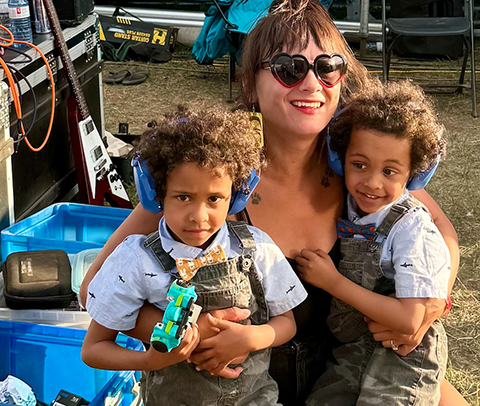 Twin boys wear earphones and their musician mom has her arms around them.