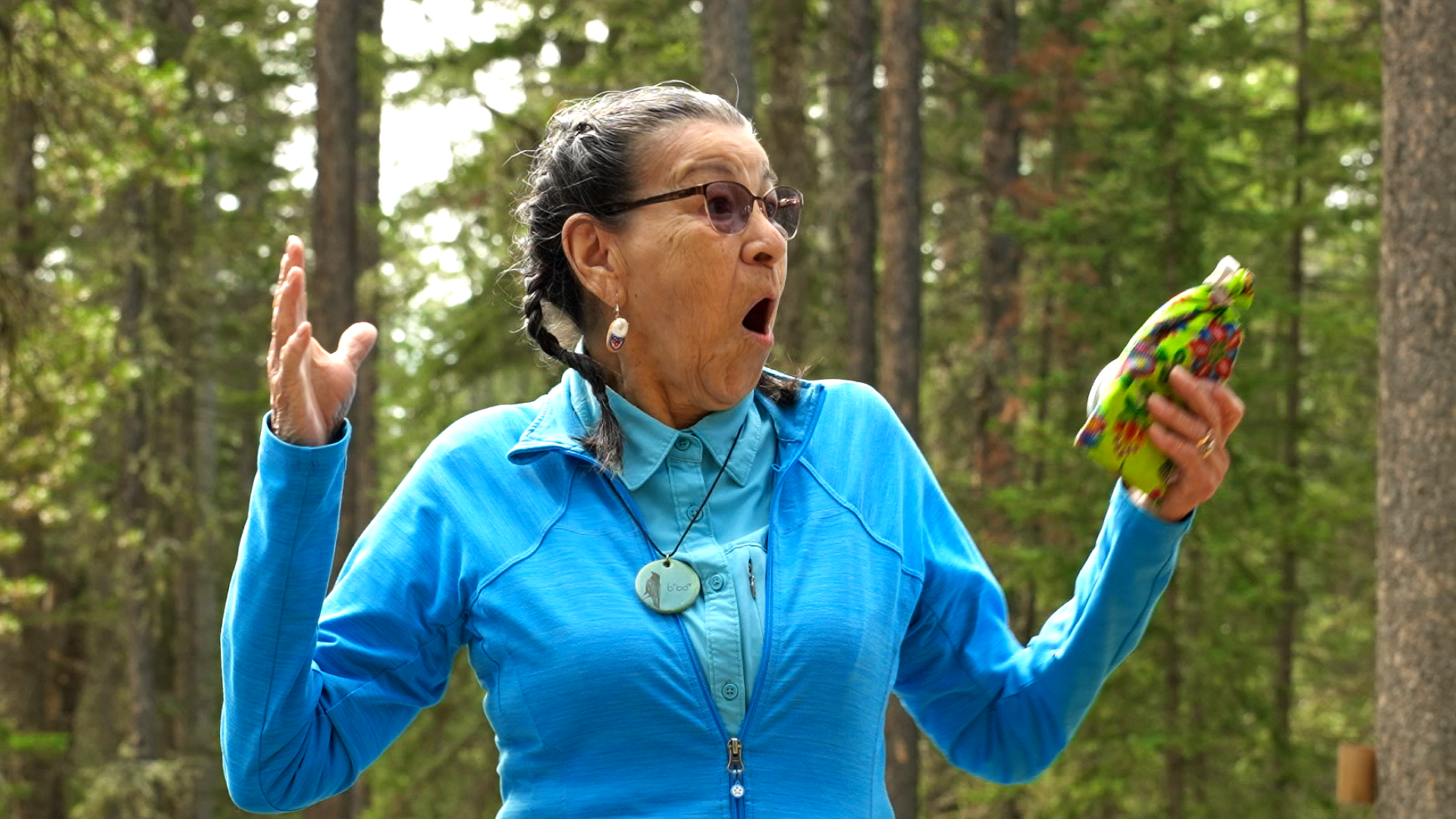 Surprise! A woman has her hands up and a look of surprise on her face. She stands amongst the trees.