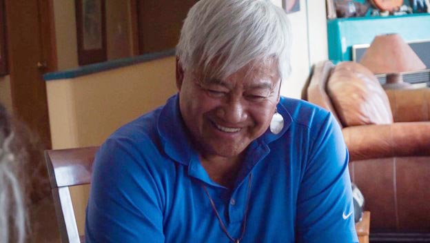 A man with grey hair smiles and looks downward. He is sitting in a kitchen chair.