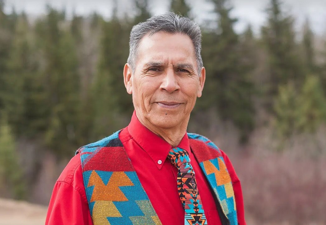 A man in a red shirt and multi-coloured vest and tie stands in front of a backdrop of trees.