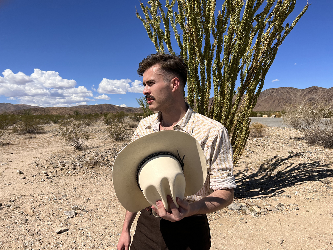 A man stands with a cowboy hat in his hands while out in a desert-like landscape.