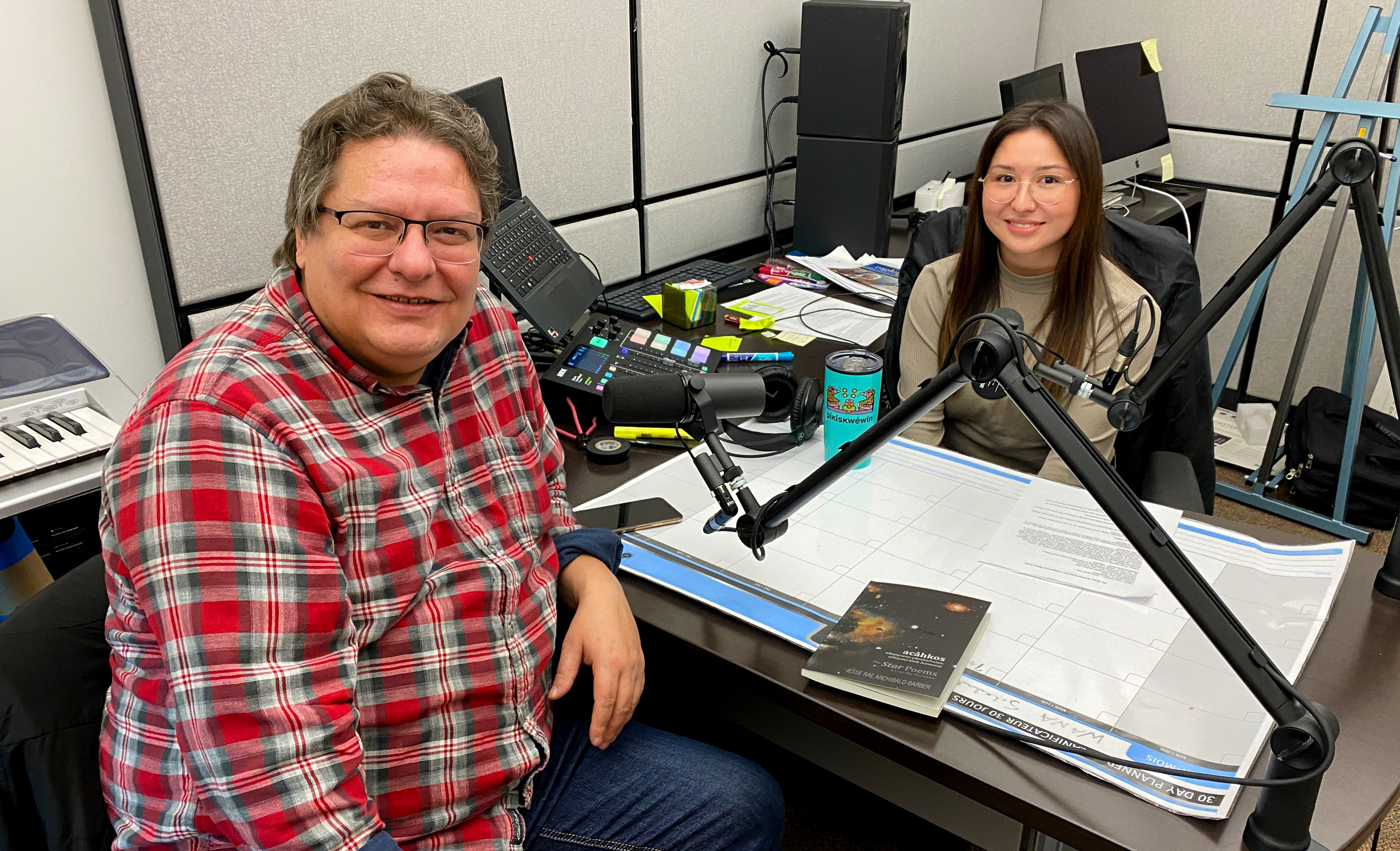 A man sits in a radio station booth with a young woman. Each has a radio microphone in from of them and notes on the desk top between them.