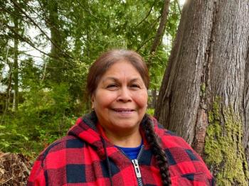 A smiling woman wears a red and black jacket while standing among trees.