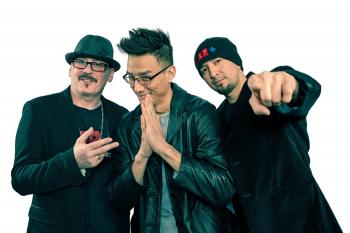 Three Indigenous men dressed in black jackets pose for the camera. One with his hand on his heart. Another with his hand in front of his chest in a prayer, and the other man pointing his finger towards the camera.