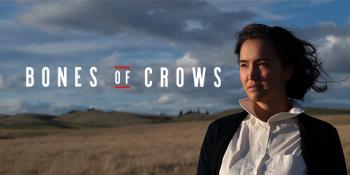 Bones of Crows poster shows a woman standing in a field, wind blowing her dark short hair. Dark clouds are gathring in the blue sky behind her.