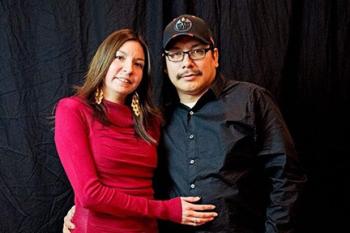 A woman in a red top stands with a man in a black shirt and ball cap, their arms around each other's waists.