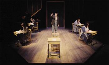 The character of a residential school student stands centre stage surrounded by students sitting at old-fashioned school desks.