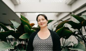 Behind a woman who stares toward the camera is a backdrop of large leafy indoor plants.