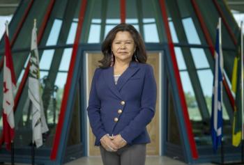 A woman stands in front of flags inside a building with a lot of windows. She is wearing a blue jacket, has her hands folded in front of her. She has dark shoulder-length hair.