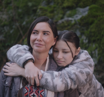 A young girl stands behind her mother wrapping her arms around the woman's shoulders.