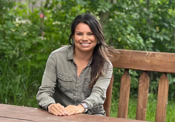 A young woman sits at on a bench behind a wooden table. It's outdoors and there is a leafy backdrop. She has her hands folded on the table in front her. She is smiling widely.