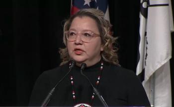 A woman speaks at a microphone. She stands in front of flags. 