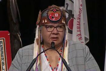 A woman in a grey suit speaks at the microphone. She wears a birchbark headband with shells on it and white fur pieces dangling from it. On it is a medallion with the AFN logo on it.