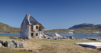 An abandoned wood-constructed home on a grassy and rock-strewn landscape is falling in on itself. The roof is caving in, and one side of house lay on the ground next to a body of water.