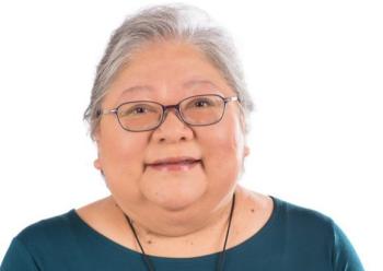 This is a photo of a woman in a teal shirt. She is grey-haired, and wears glasses. She smiles towards the camera. 