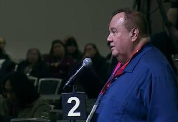 A man in a blue shirt speaks at a microphone with the number 2 on it.