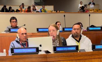 Two men and one woman sit at microphones. In from of them is a plaque identifying them as being from an Indigenous organization