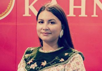 A woman in a traditional cotton dress with beaded collar stands against a red background smiling slightly.