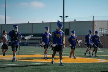 Lacrosse players walk across the playing field. They wear purple jerseys. 