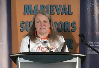 A woman stands at the podium with a panel behind her that reads Marieval Indian Residential School