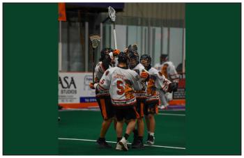 A jubilant group of lacrosse players meet and hug in a box arena.