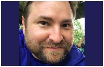 A man in a blue T-shirt looks directly into the camera. He has a beard and a mustache.