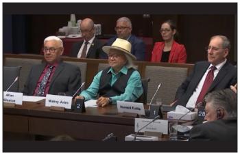 Three men sit at a desk with three people sitting behind them. They address the Senate of Canada.