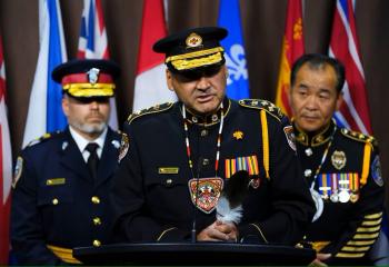 Three police men in full uniform, two wearing hats, stand in front of a bank of flags. One stands before a microphone to speak.