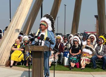 A man in a blue suit and a chief's headdress stands in front of a group of people who are seated. He is at a podium and speaking into a microphone.