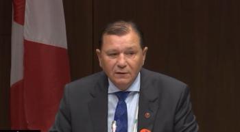 A man in a suit speaks. Behind him is a Canadian flag.