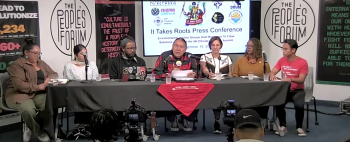 A group of people sit at a table with microphones on it. There are banners that read "The Peoples' Forum" on each end and behind the group.