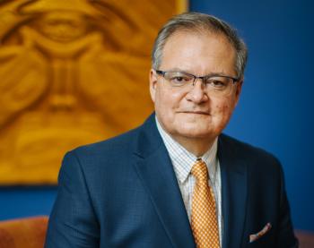 A man looks toward the camera. He is pictured in front of a large wood carving on the wall behind him.