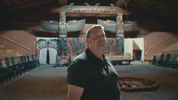 In the background are huge winged totem poles on each end of a painted cedar wall screen. Painted ceremonial curtains can be seen as are other cedar carved objects. A stepped wooden seating area flows upwards on either side of a dirt floor in the large building. In the centre is a place for a ceremonial fire. A man stands in the foreground looking upwards.