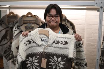 A woman with short dark hair and wearing a Cowichan sweater holds up a Cowichan sweater with a snowflake design in front of her. Behind her hang Cowichan sweaters.