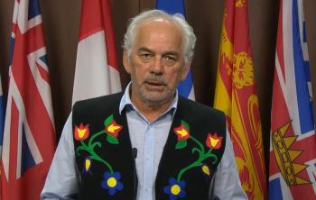 A man stands at a microphone in front of a row of flags.