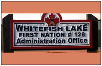 Two wooden posts hold up a sign with a red maple leaf with a tipi overlaid on it. The sign reads WHITEFISH LAKE FIRST NATIONS #128 Administration Office