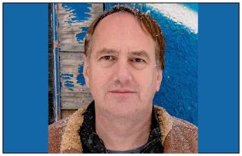 A man is photographed in front of a building with peeling blue paint on its walls.