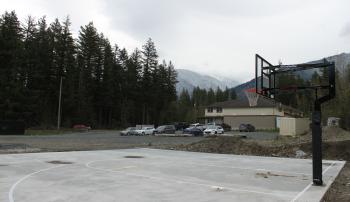 A mist gathers over far away mountains in the background. A cement pad has been marked out for a basketball court. A hoop and backboard are seen.