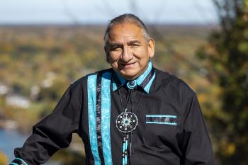 A man in a black shirt with turquiose trimming stands among the trees smiling towards the camera.