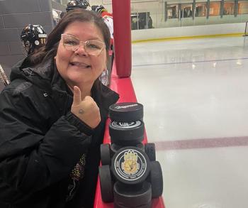 A woman stands in the players box in a ice rink. She raises her thumb as she smiles at the camera. A stack of pucks sit on top of the boards in front of her.