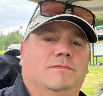 A man looks into the camera. He wears a cap with a pair of sunglasses perched on it.