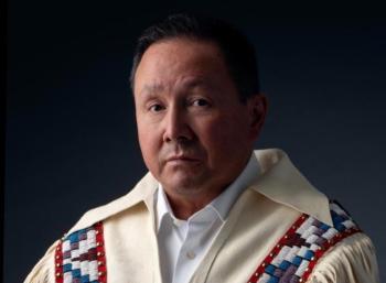 A man with a white fringed jacket with beading over the shoulders looks into the camera without smiling.