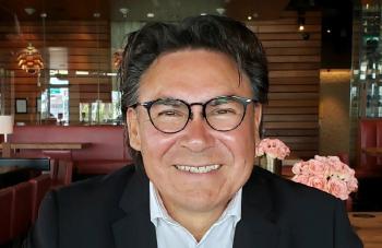 A man sits at a table in a restaurant. Flower arrangements are on tables. He smiles into the camera.