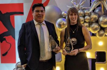 Two people, a man and a woman, are holding awards. In the background is a set made up of banners and balloons.