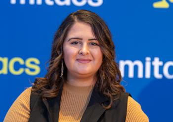 A young woman with long dark hair wears a mustard coloured top under a black vest. She stands in front of a blue screen with the word "mitacs" printed in white on it.