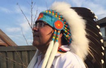 A man stands outside in a yard. He wears a chiefs feather headdress.