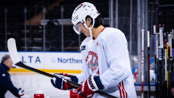 A man wearing a Washington Capitals hockey uniform is standing in the players' bench smiling and looking at the stick in his hands.